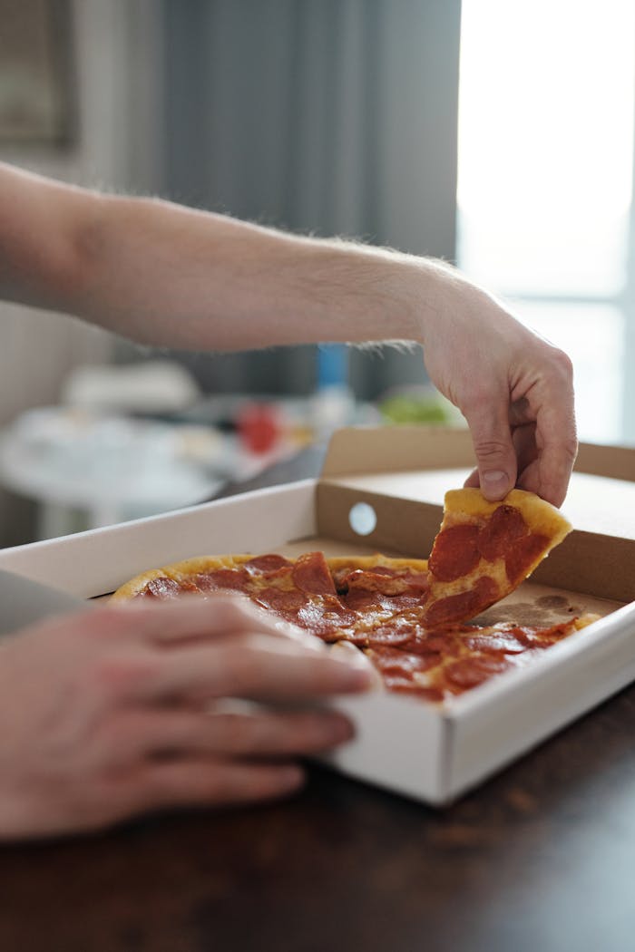 Close-up of hands picking a slice of pepperoni pizza from a box, showcasing a delicious and casual dining moment.